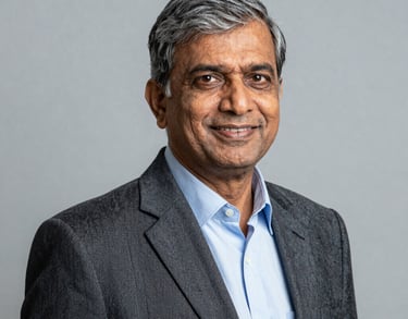 A professional studio portrait of a senior South Asian male educator wearing a formal blazer, smiling confidently. The lighting is professional and the background is a solid light gray.