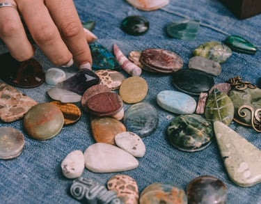 A hand sorts through various polished gemstones and healing crystals on blue denim fabric.