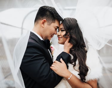 A smiling bride and groom embracing under a flowing white wedding veil during a romantic outdoor ceremony.