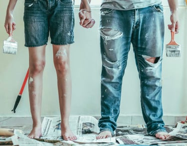 A couple in paint-splattered jeans holding hands and brushes during a DIY home renovation.
