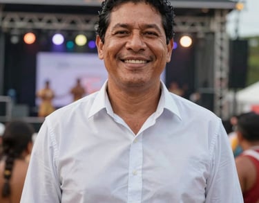A professional South American / Brazilian Amazonian outdoor portrait cropped into a square shape of a male cultural producer in Santa Bárbara do Pará. Smiling warmly, wearing a crisp light shirt near a festive stage.
