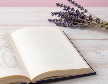 A detail of an open book on a pale wood table next to a sprig of dried lavender, soft pearl pink background, Southern European / Italian office.
