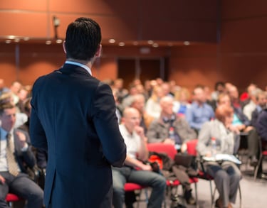 Professional public speaker in a business suit giving a presentation to a large audience at a conference.