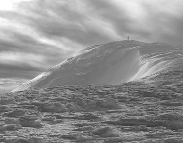 Black and white landscape of a lone hiker standing on a snowy mountain peak under a dramatic sky.