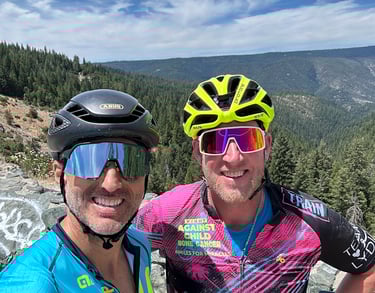 Two cyclists (men) posing for a picture at the top of a mountain range