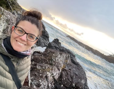 The founder of Alignment Era Consulting smiling on a rocky coast at sunset with ocean waves.