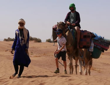 A boy leads a man riding a camel on a Sahara desert trek, while a nomad guide walks in front