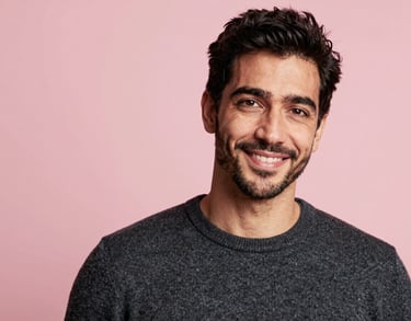 A headshot of an International / English-speaking professional man with a friendly expression, wearing a dark grey sweater, standing against a soft light pink background.