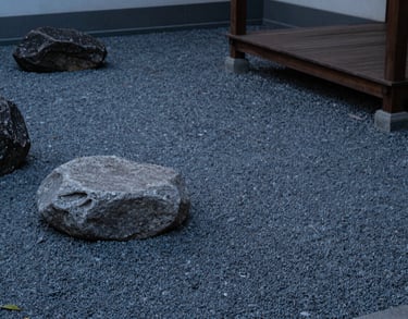 A photograph of a serene zen garden corner with smooth grey stones and a small wooden structure, captured in soft evening light.