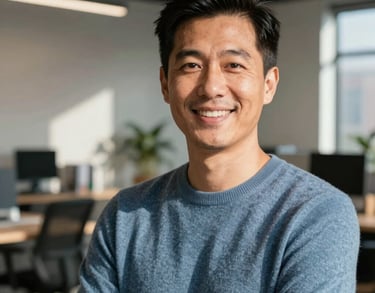 A professional headshot of a person in a muted blue sweater, smiling confidently in a sunlit modern workspace. North American / US urban office setting.