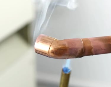 Professional plumber soldering a copper pipe elbow joint with a blue propane torch flame and rising smoke.