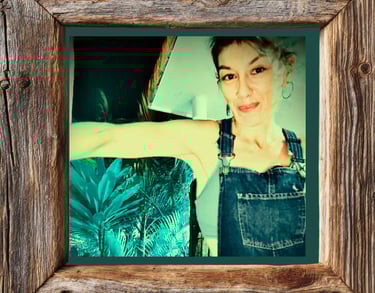 Smiling woman in denim overalls framed by rustic reclaimed wood with tropical plants in background.