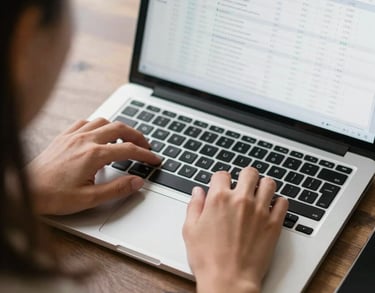 An over-the-shoulder shot of a person typing on a sleek laptop with a clean spreadsheet visible on the screen. Natural light, professional, North American / US.