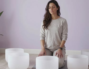 A professional portrait of a sound healer standing next to large crystal bowls in a North American holistic center. The room is decorated in soft white and pale lavender tones.