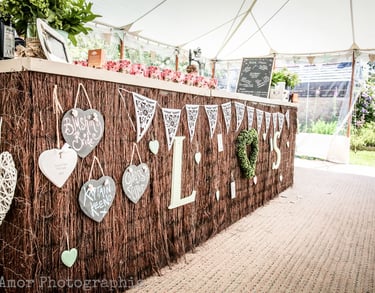 Rustic outdoor wedding bar decorated with heart signs and lace bunting under a white marquee tent.