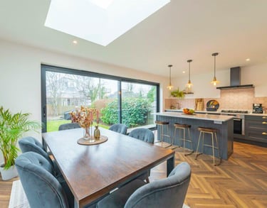 Modern open plan kitchen and dining room with herringbone floors, large skylight, and sliding glass doors.