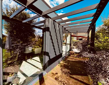 Modern garden pergola with a wooden deck, glass walls, and a hanging chair in a lush backyard.