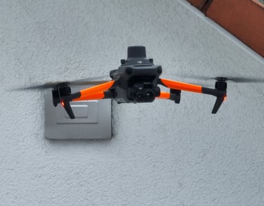 A DJI Mavic 3 Enterprise drone with orange arms hovering near a building wall for a roof inspection.