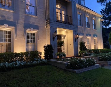 Luxury white brick home at dusk featuring warm exterior landscape lighting and manicured gardens.