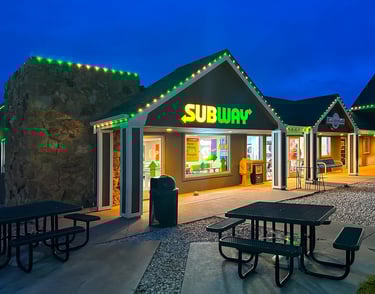A Subway sandwich restaurant storefront at night with green roof lights and outdoor patio seating.