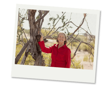 Smiling senior woman in a vibrant red shirt posing outdoors by a desert tree.