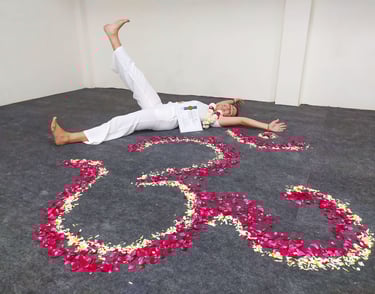 Eva lying on the ground next to a flower mandala, smiling happily while holding her Yoga Teacher Training diploma in Rishikes