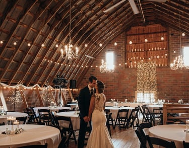 Newlywed couple dancing in a rustic barn wedding venue with string lights and chandeliers.