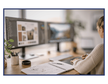 A graphic designer works on dual monitors using a digital drawing tablet and notebook at a wooden desk.