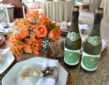"Mesa de casamento decorada com arranjo floral de rosas laranjas e gipsofila, garrafas de Lambrusco 