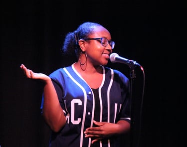 a Black woman in a navy baseball jersey and glasses speaking into a microphone