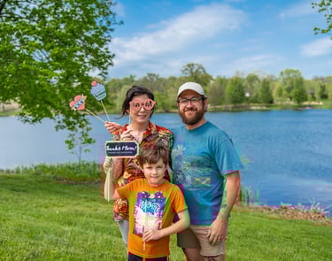 Staged photo of a couple with a young child in front of a lake, holding up paper props