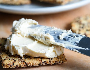 Cuchillo untando queso crema vegano hecho de macadamia sobre una galleta cracker, mostrando textura suave y blanca
