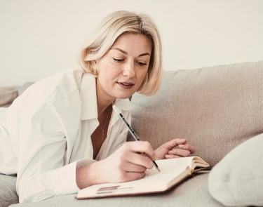 Blonde woman lying on a grey sofa writing in her journal or planner with a pen.
