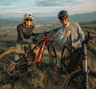 Two mountain bikers with helmets and e-bikes posing on a scenic hilltop trail at sunset.