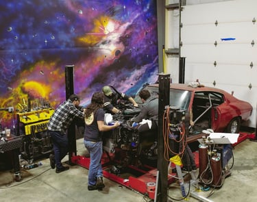 Mechanics working on a classic car engine in a garage with a cosmic mural.