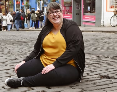 Laura is laughing at the camera sat on a cobbled street with her legs curled up under her