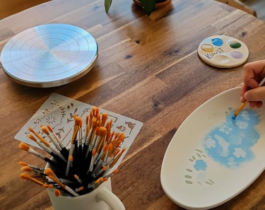 An artist hand paints blue flowers on a ceramic plate using a fine brush at a pottery studio table.