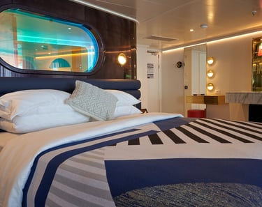Modern luxury cruise ship cabin featuring a plush bed with geometric bedding and an oval porthole window.