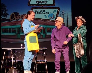 Actors in colorful tracksuits perform a comedic theater scene in front of a retro diner backdrop.