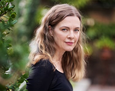 A beautiful middle-aged woman with long curly hair and a black shirt standing in a garden and looking at the camera