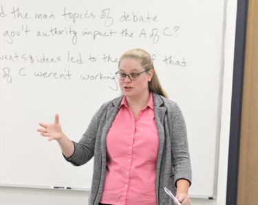 Dr. Hooton in front of a white board at the front of a classroom, teaching.