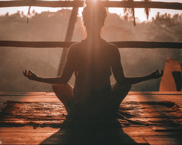 Silhouette of a woman practicing morning yoga meditation on a wooden deck during a tropical sunrise.