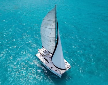 An aerial view of a white sailing catamaran cruising through clear turquoise tropical ocean water.