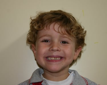 A smiling young boy with curly blonde hair showing his teeth in a happy portrait.