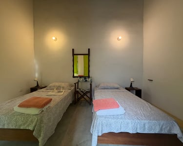A minimalist guest room featuring two twin beds, bedside lamps, and a folding chair below a wall mirror.