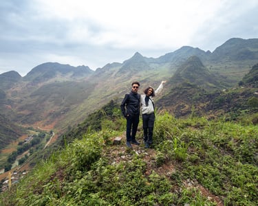 A couple standing on a grassy mountain edge posing for the camera. in the background are the beautiful Ha Giang mountains.