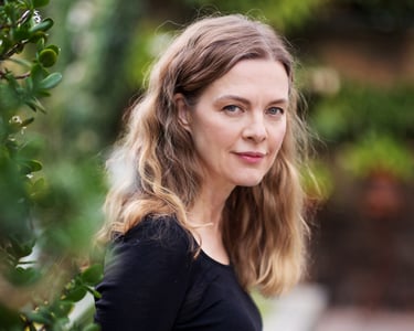 A beautiful middle-aged woman with long curly hair and a black shirt standing in a garden and looking at the camera