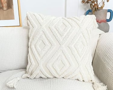 White boho tufted throw pillow with textured diamond pattern and corner tassels on a neutral sofa.