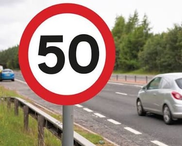 A circular red and white 50 mph speed limit sign on a roadside with cars passing by.