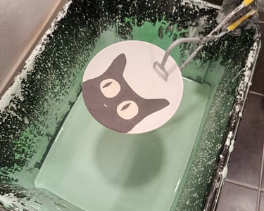 A ceramic plate with a hand-painted black cat being dipped into a bucket of green pottery glaze.
